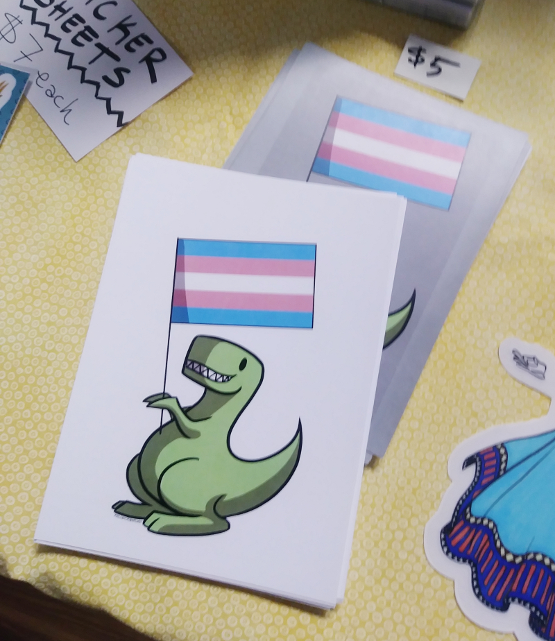 New Mr. Dino Postcards! A portion of sales of these will go towards Margie's Hope in Lakewood, OH. On the postcard is a dinosaur holding a transgender pride flag.