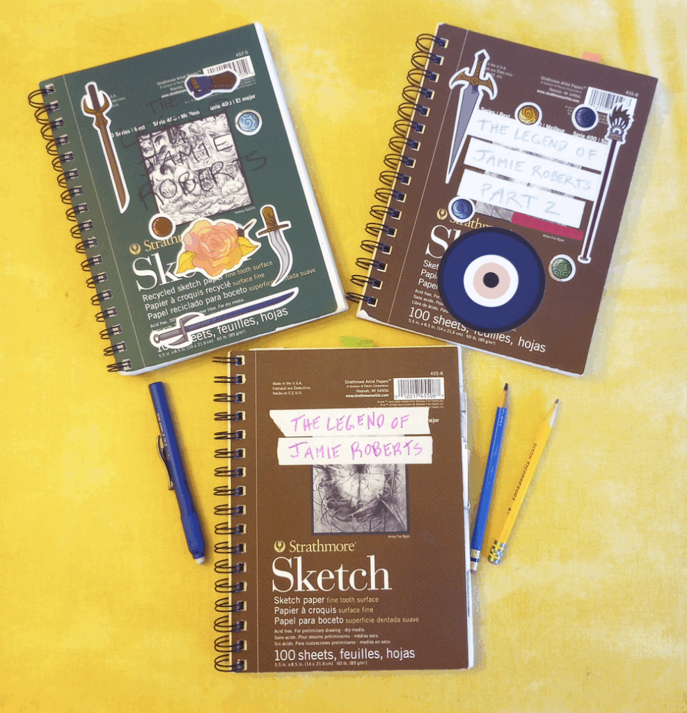 three sketchbooks, their covers covered in notes and stickers, are laid on a yellow tablecloth