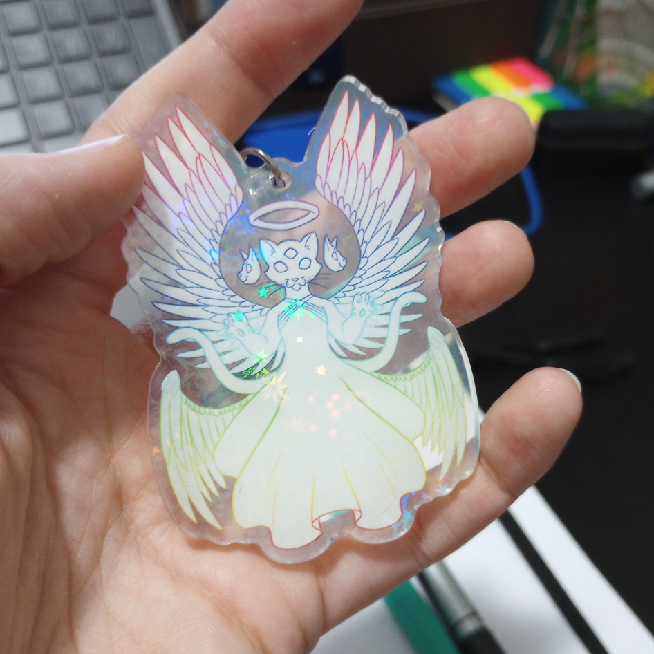 a pale hand holds a holographic keychain. It depicts a biblically accurate cat angel.
