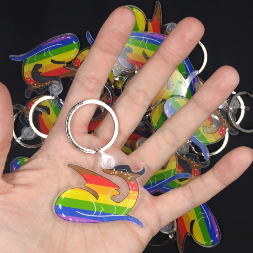 a pale hand with long fingers holds a keychain. The keychain is a baby dragon painted in the colors of the Philadelphia LGBTQ+ Pride flag.