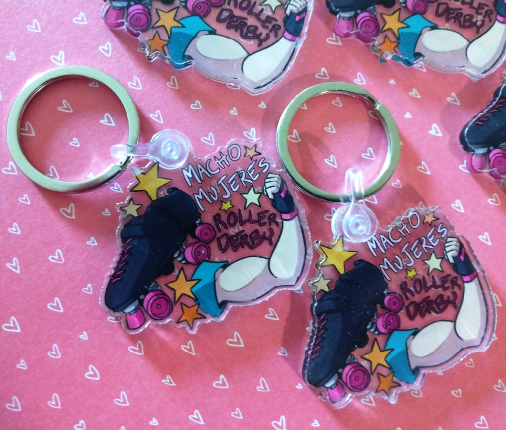 a picture of two keychains, both identical. They show an arm flexing and a roller skate with pink wheels. Text and stars decorate both, saying "Macho Mujeres Roller Derby"